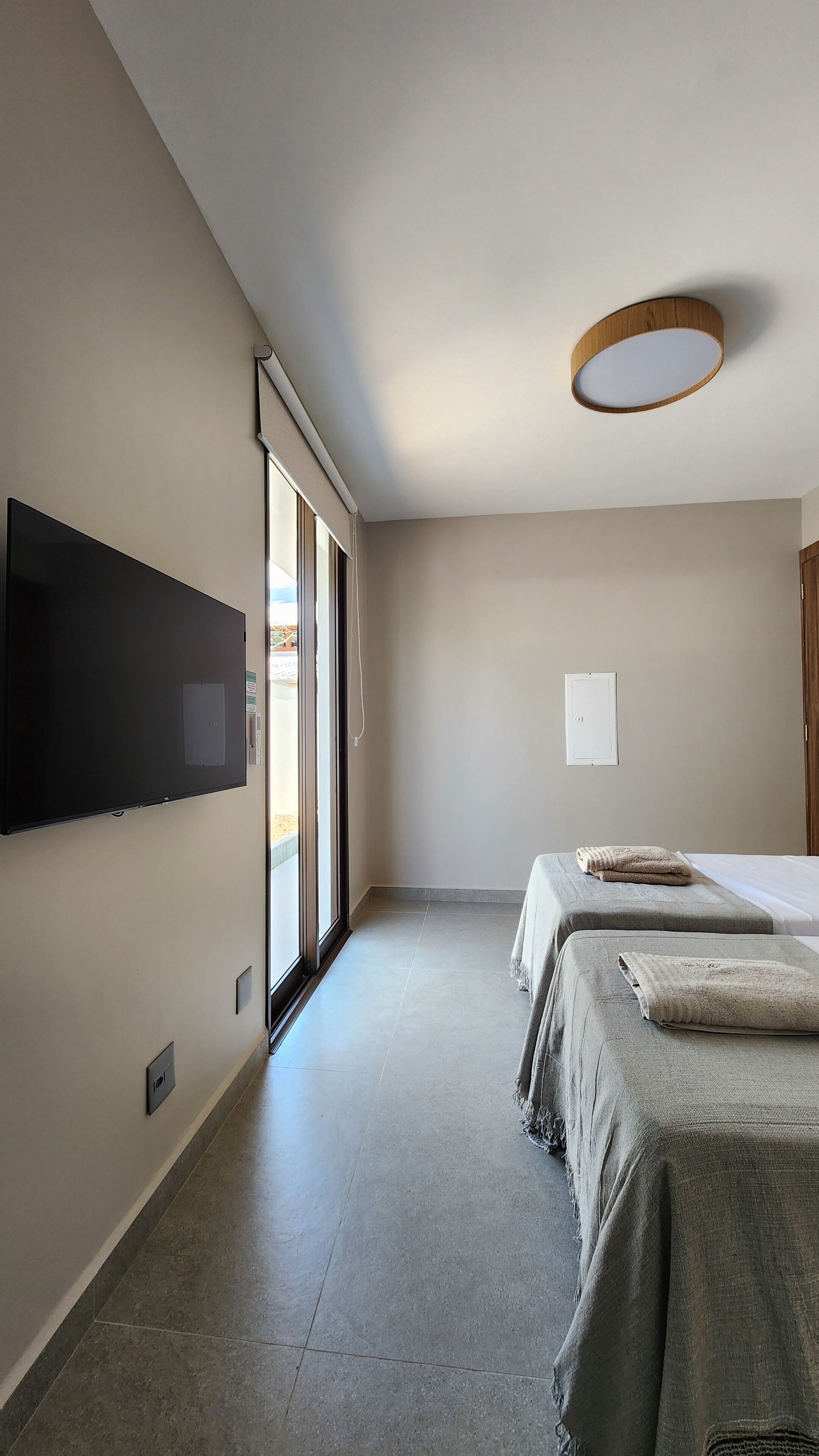 Una habitación de hotel moderna con un televisor de pantalla plana grande montado en la pared, una cama con manta gris y una lámpara de techo.