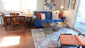 A living room with a blue sofa, a coffee table, and a dining area with a wooden table and chairs. - Sweet cottage nestled in scenic Drummer's Cove (CS0052) (Wellfleet)