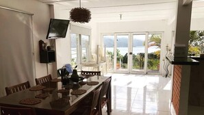 Comedor con una mesa dispuesta para cuatro, un televisor montado en la pared y vista al mar a través de amplias puertas correderas.
