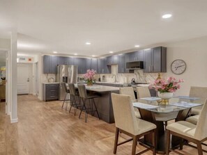 A modern kitchen with grey cabinets, a stainless steel refrigerator, and a dining area with a glass table and chairs. - Longboat Key Shore Beach View (Longboat Key)