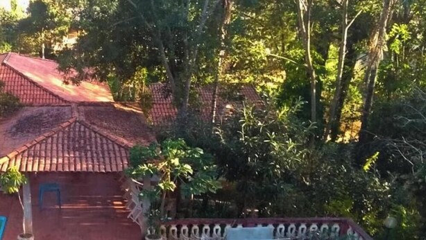 A swimming pool with a tiled deck, surrounded by greenery and a small building with a red roof. - Pousada das Cores Cocais (Cocais)