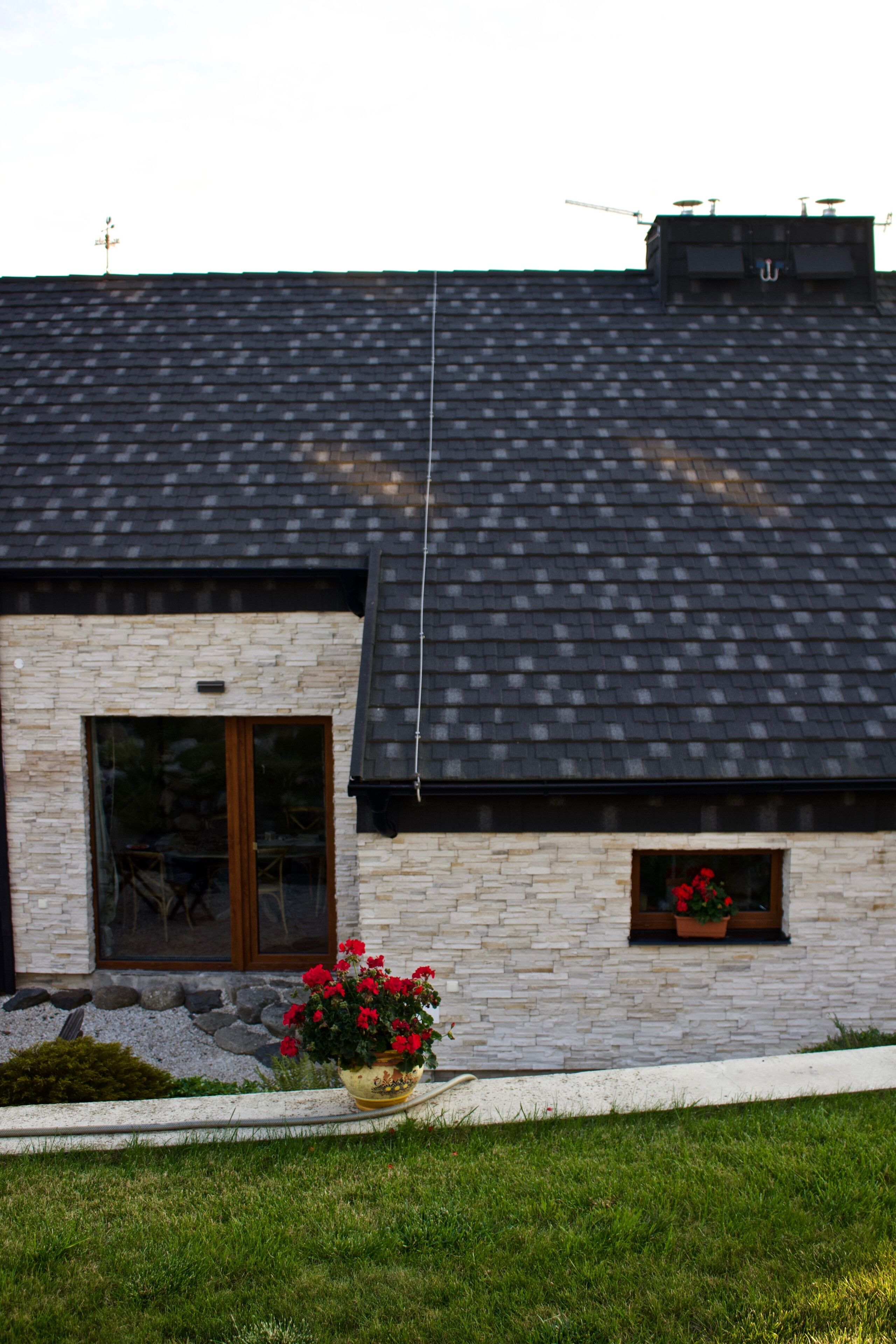 A modern house with a stone facade, a dark tiled roof, and a small window with flowers.