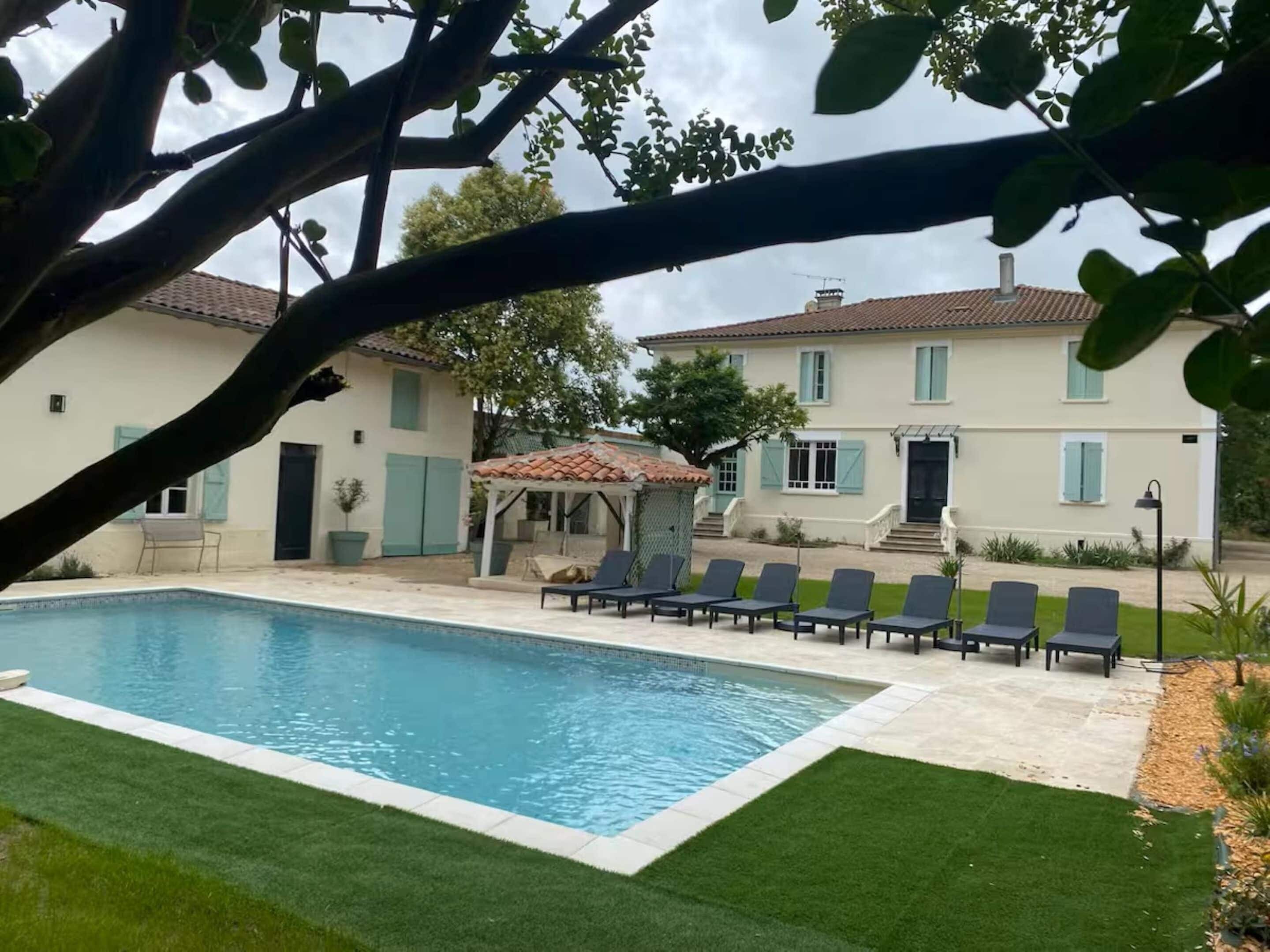 A swimming pool with lounge chairs, a gazebo, and a house with a tiled roof and shutters.