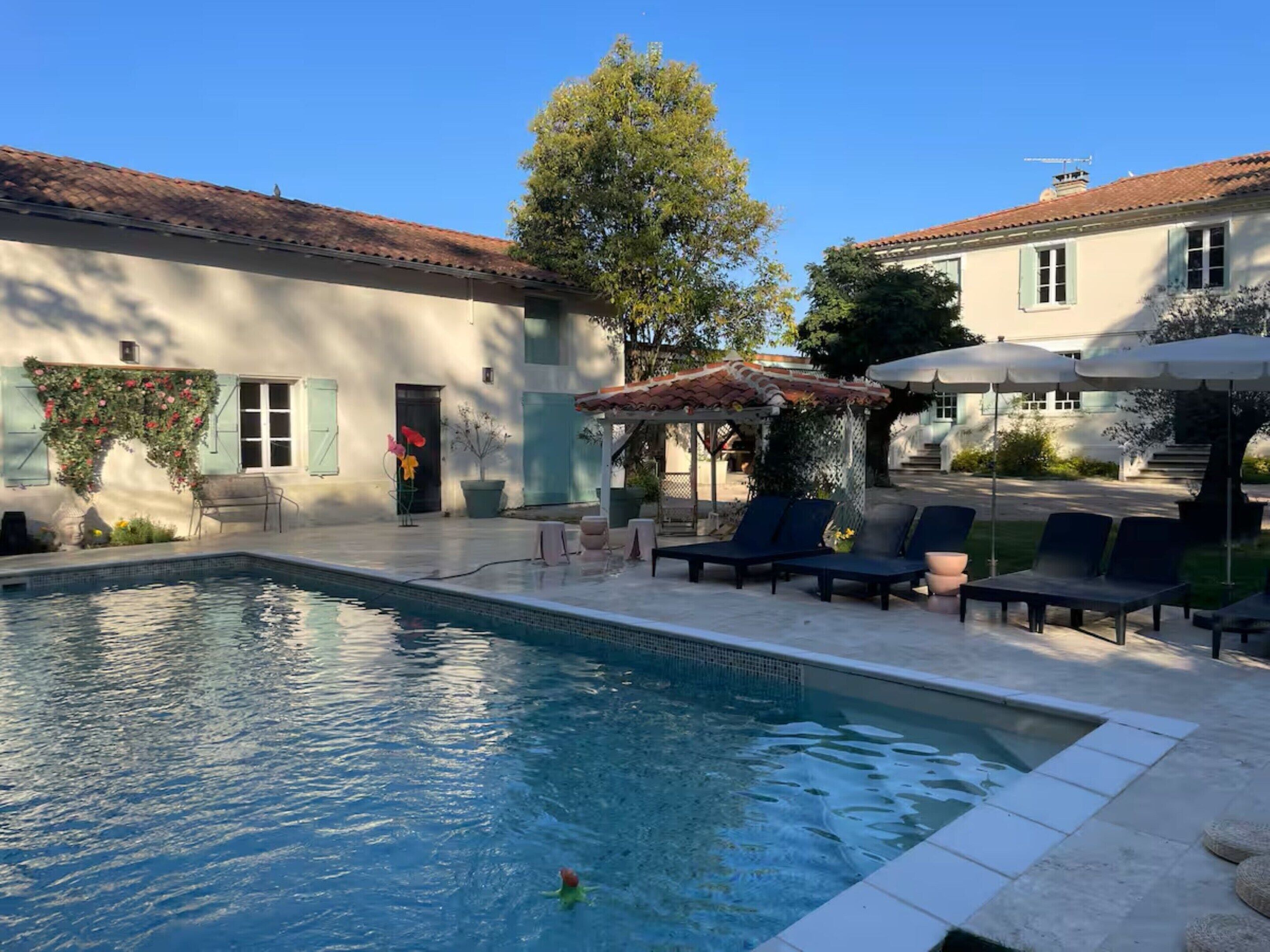 Une piscine entourée de chaises longues et de parasols, le tout bordé d’un bâtiment doté d’un toit en tuiles et d’un jardin.