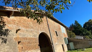 A brick building with a large arched entrance, a small window with shutters, and a grassy area in front. - The Oasis Gardens, Beautiful House (Barry-d'Islemade)