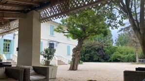 A covered outdoor seating area with wicker furniture, a tree, and a building with blue shutters in the background. - The Oasis Gardens, Beautiful House (Barry-d'Islemade)