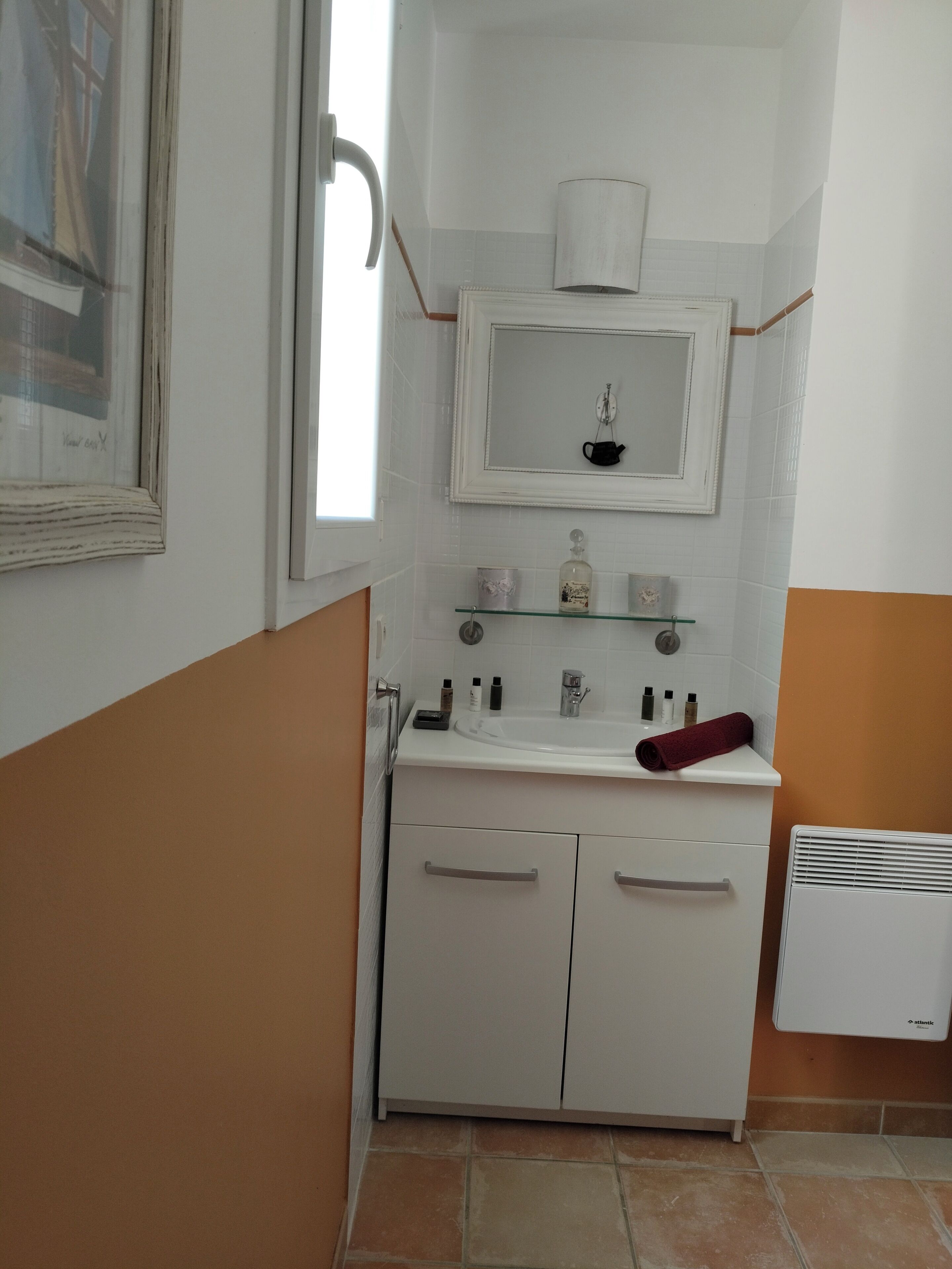 Une salle de bain avec un lavabo blanc, un miroir et un porte-serviettes. Il y a une fenêtre donnant sur un immeuble, et un radiateur blanc posé sur le sol.