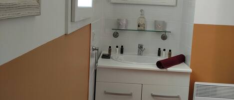 Une salle de bain avec un lavabo blanc, un miroir et un porte-serviettes. Il y a une fenêtre donnant sur un immeuble, et un radiateur blanc posé sur le sol.