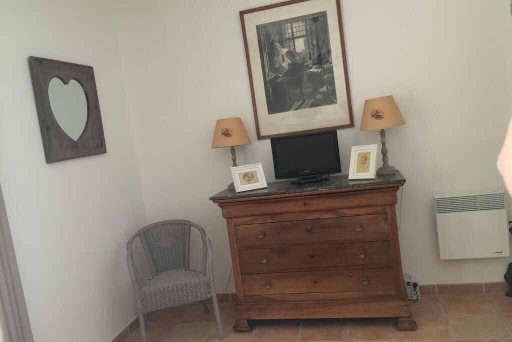 A room with a wooden dresser, two framed pictures, a television, a lamp, a chair, and a wall-mounted heater.