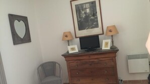 A room with a wooden dresser, two framed pictures, a television, a lamp, a chair, and a wall-mounted heater.