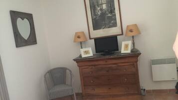 A room with a wooden dresser, two framed pictures, a television, a lamp, a chair, and a wall-mounted heater.