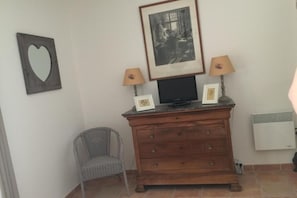 Une pièce comprenant une commode en bois, deux tableaux encadrés, un téléviseur, une lampe, une chaise et un radiateur fixé au mur.