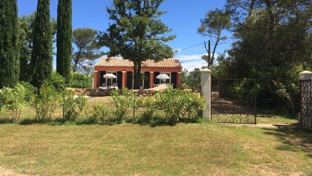 A building with a tiled roof and columns, surrounded by a fenced garden with various plants and trees.