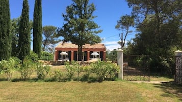 A building with a tiled roof and columns, surrounded by a fenced garden with various plants and trees.