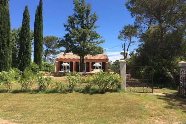 Un bâtiment doté d’un toit en tuiles et de colonnes, entouré d’un jardin clos où poussent diverses plantes et arbres.
