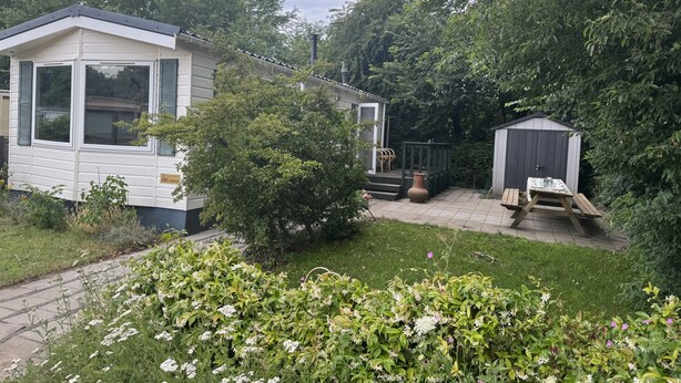 Een stacaravan met een klein terras en een tuinhuis in een weelderige tuin.