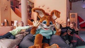 Een zacht pluche konijn knuffel zit op een bed met kussens, omringd door kinderen.