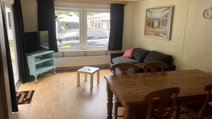 Een woonkamer met een houten tafel, twee stoelen, een bank, een televisie op een standaard en een raam met gordijnen.