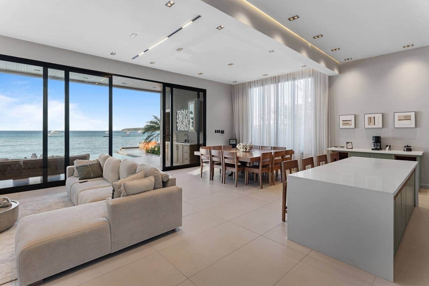 A modern living room with a large window overlooking the sea, a dining area with a central island, and a kitchen with a view.
