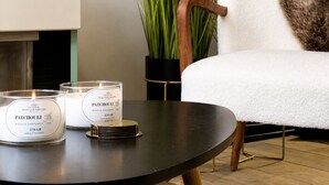 Um quarto com uma mesa redonda preta, duas velas rotuladas 'PACHOULI', uma cadeira branca com um cobertor de pele e uma planta verde alta em um vaso preto.