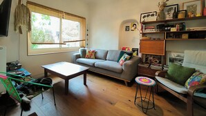 Un salon chaleureux avec un canapé gris, une table basse en bois, un fauteuil vert et un tabouret coloré. Un téléviseur est fixé au mur, et une bibliothèque contient divers objets.