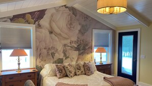 Ein Schlafzimmer mit einem großen Bett, zwei Nachttischen, einem Sessel, einer Tür und einem Fenster mit Jalousien.