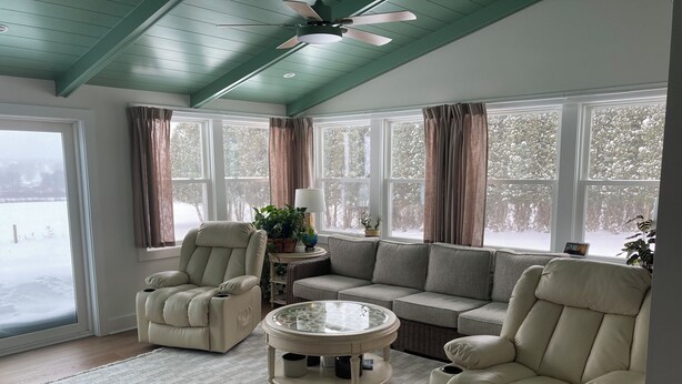 Ein gemütliches Wohnzimmer mit einem beigefarbenen Sofa, zwei Sesseln, einem Glastisch und einem Deckenventilator. Große Fenster bieten Blick auf eine verschneite Landschaft.