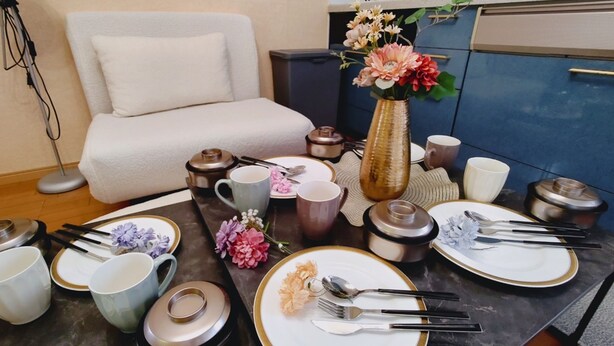 A neatly arranged dining table with plates, cutlery, and a vase of flowers, set against a backdrop of a sofa and cabinets.