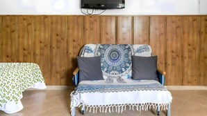 A room with a wooden panel wall, a mounted television, a patterned sofa with two pillows, and a bed with a green and white patterned bedspread. - Peaceful Apartment Close to the Coast (Conil de la Frontera)