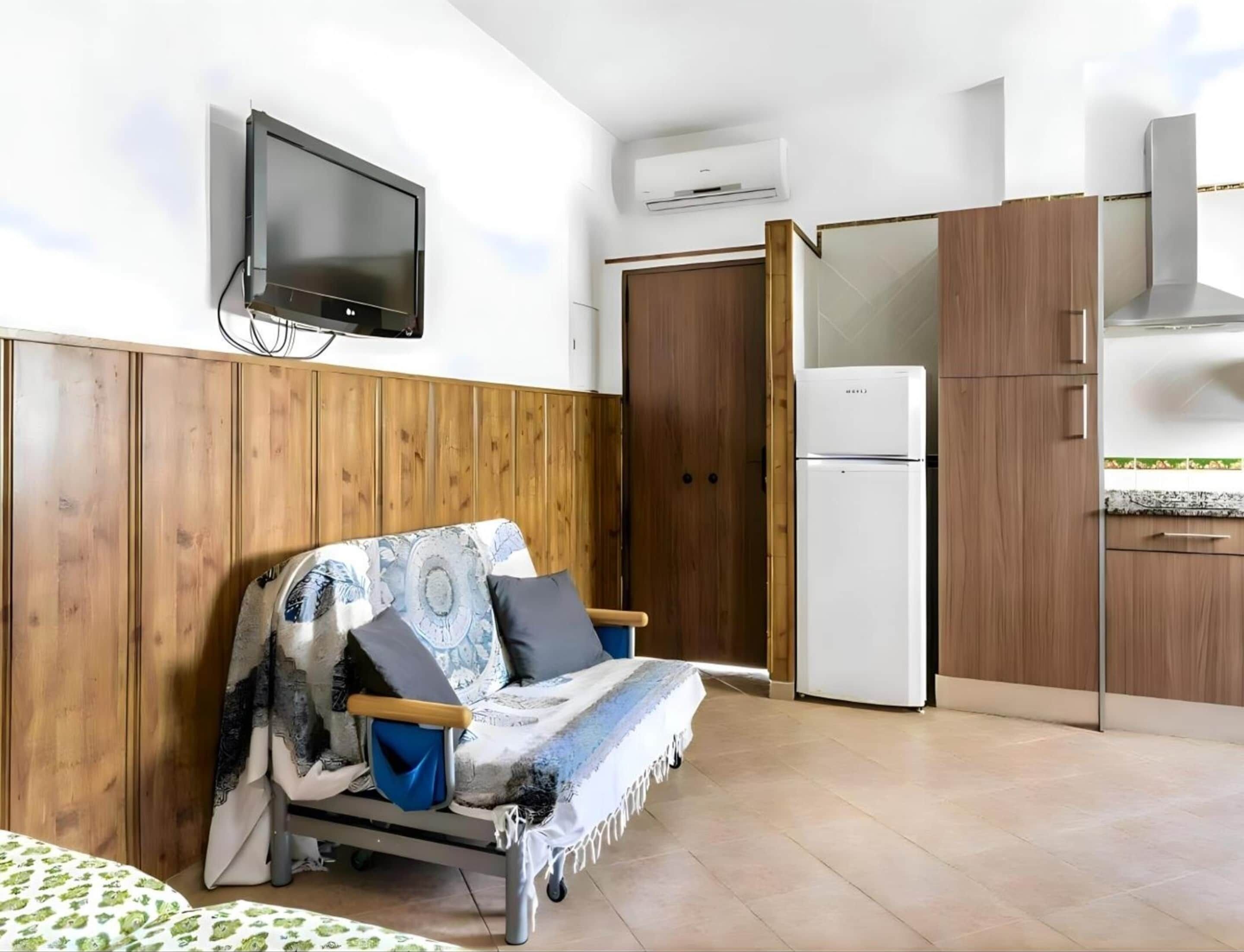 A compact room with a sofa, a flat-screen TV, a refrigerator, wooden cabinets, and a tiled floor.
