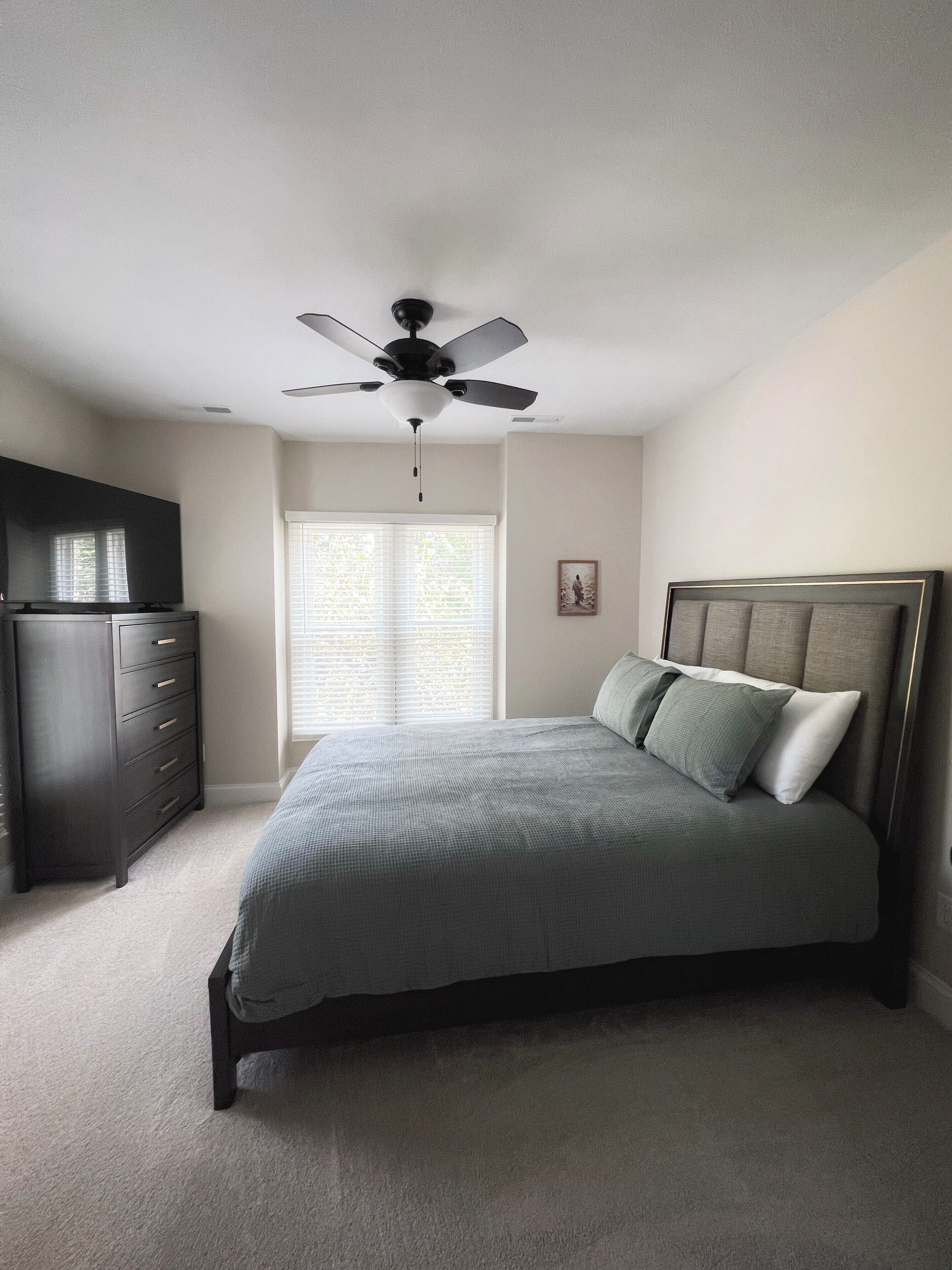 A bedroom with a bed, a dresser, a ceiling fan, and a window with blinds.