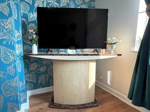 A modern living room with a flat-screen TV on a wooden stand, a small white vase with flowers, and a blue floral wallpaper.
