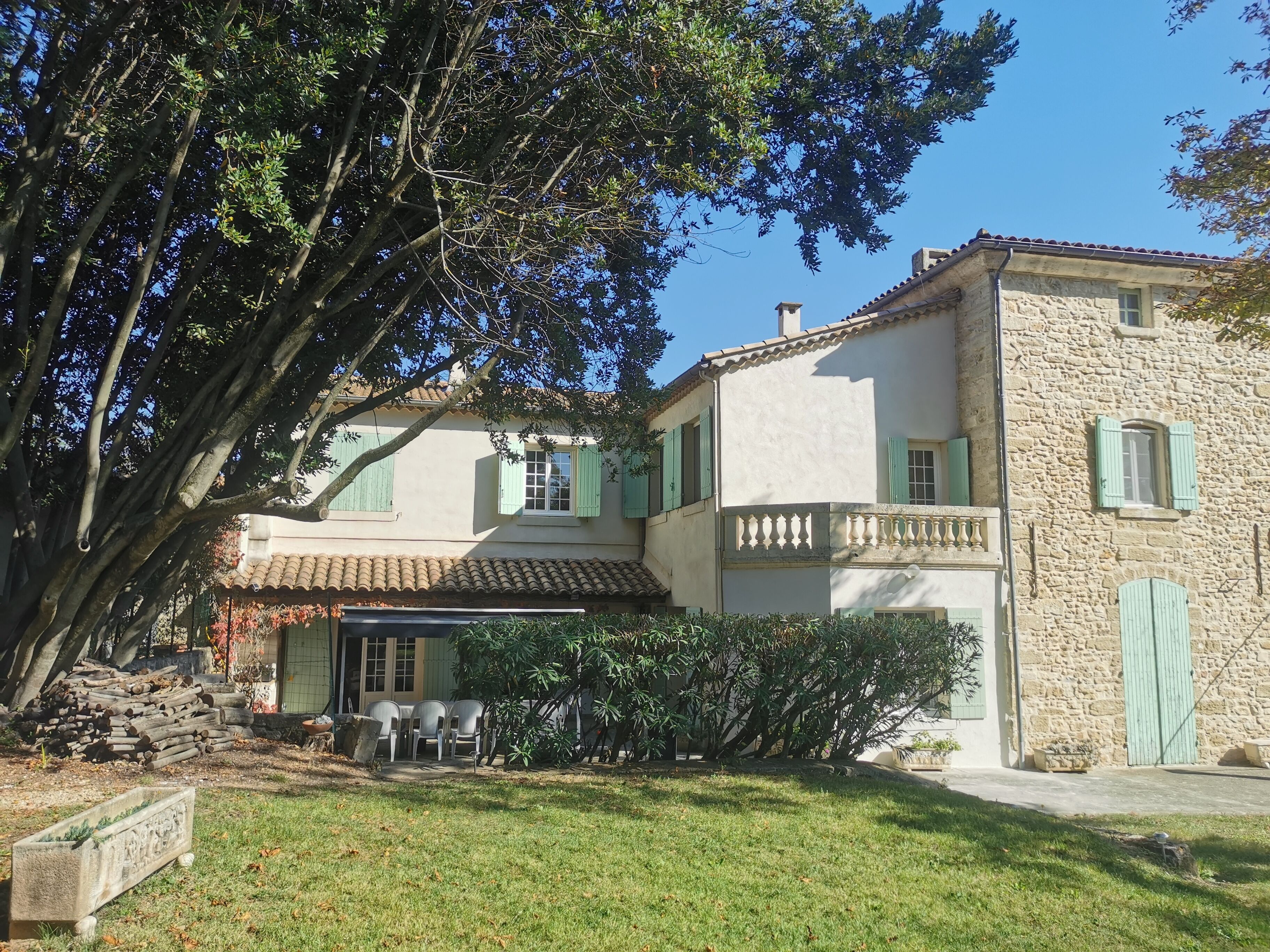 A two-story stone building with a tiled roof, green shutters, and a covered patio area with outdoor seating.