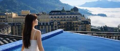 Una persona sentada al borde de una piscina, con vistas a montañas y un edificio al fondo.