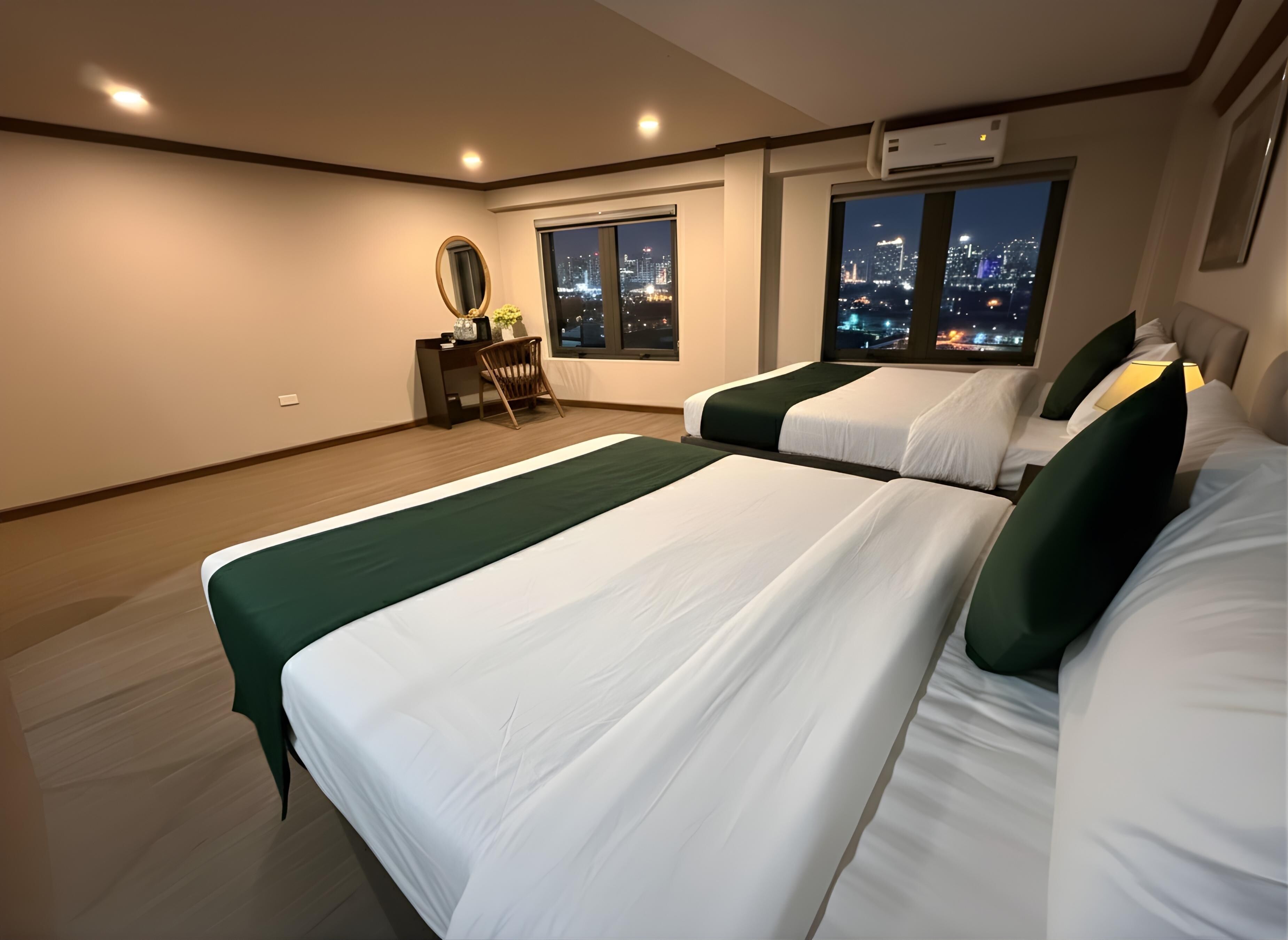 Ein Hotelzimmer mit zwei Betten, einem Schreibtisch, einem Stuhl, einem Spiegel und Blick auf die beleuchtete Stadt.