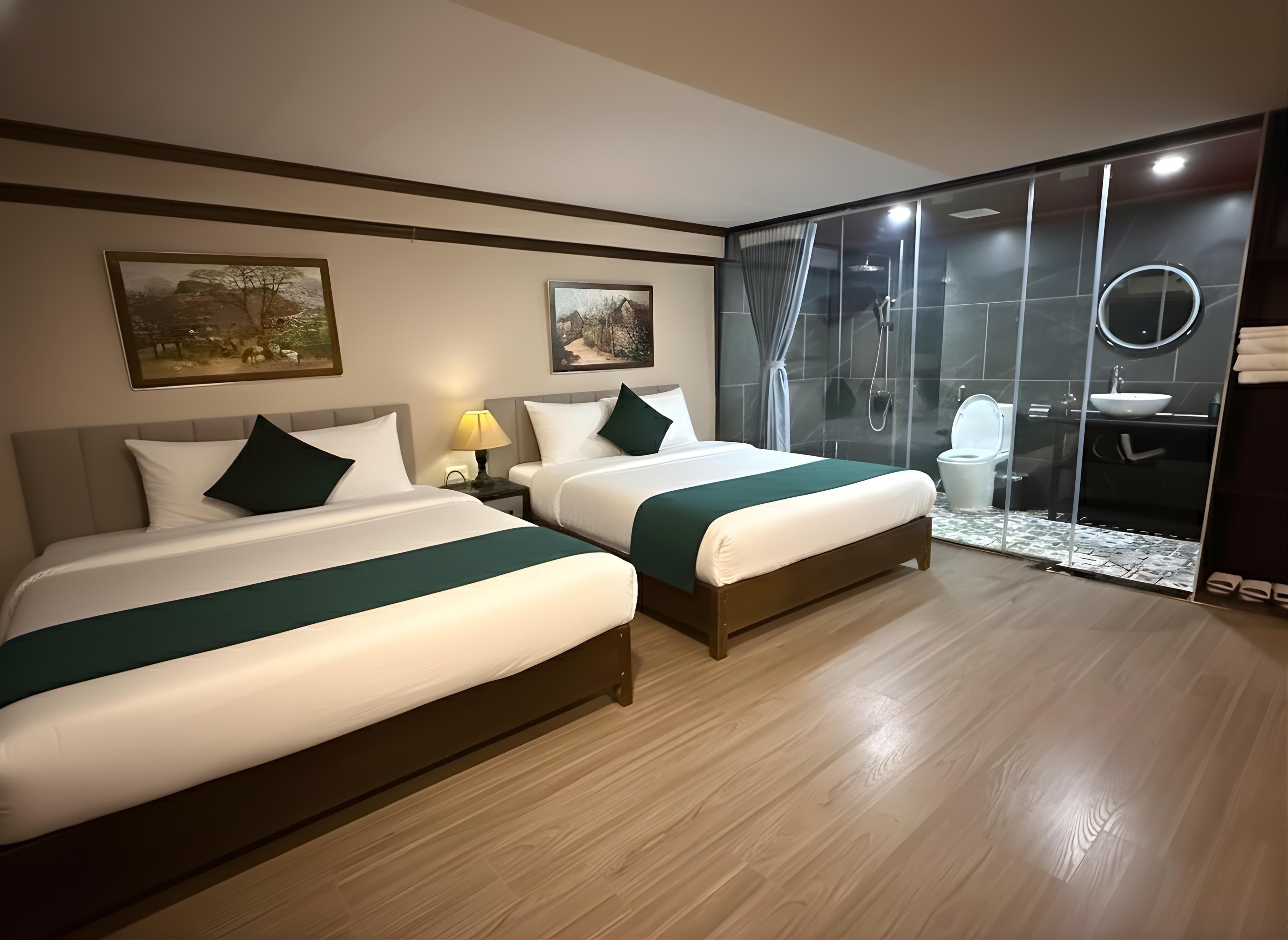 Habitación de hotel con dos camas, zona de ducha con mampara de cristal, lavamanos y inodoro.