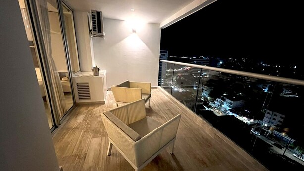 Un balcon donnant sur une vue de la ville la nuit, un petit coin salon avec une table et une fenêtre avec des rideaux.