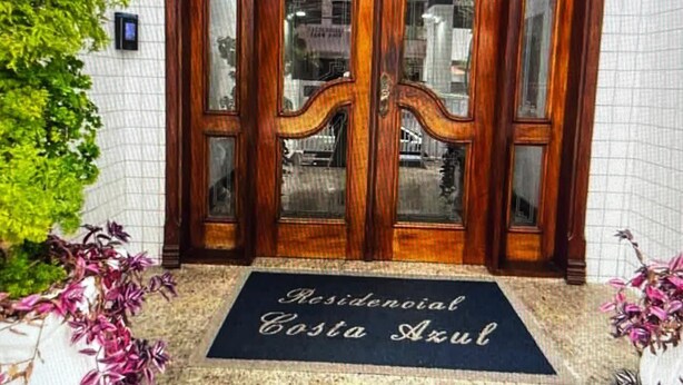 A wooden double door with glass panels, a doormat with "Residencial Costa Azul" text, and a tiled wall with a plant to the left. - FOR RENT!!! "Apartment in Praia Grande, Canto do Forte" (Praia Grande)