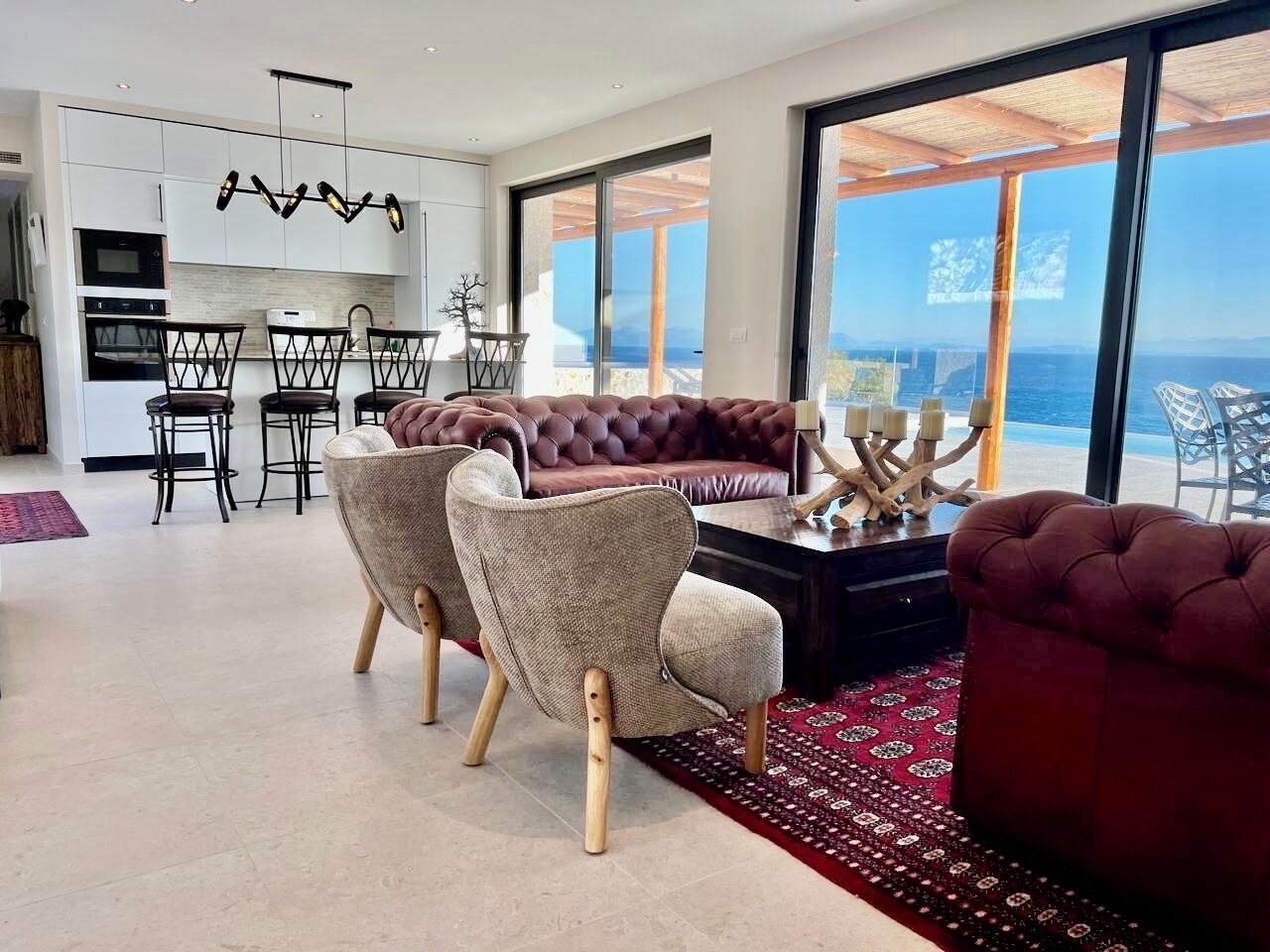 A modern living room with a large window, a sofa, two armchairs, and a dining area with a kitchen in the background.