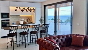 Una cuina moderna amb una illa central, tamborets i vista al mar a través de grans finestres.
