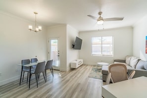 Ein modernes Wohnzimmer mit Essbereich, Deckenventilator, Flachbildfernseher und großem Fenster mit Jalousien.