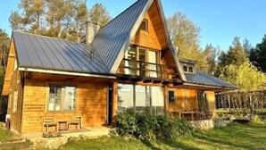 A wooden cabin with a sloped roof and large windows, surrounded by a grassy yard and trees.