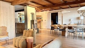 A spacious living area with a wooden ceiling, a sofa, a dining table with chairs, and a kitchen in the background.