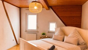 A bedroom with a sloped ceiling, a bed with a headboard, two windows, a radiator, and a hanging light fixture. - Odw Homes – Charming Home in Gorxheimertal (Gorxheimertal)