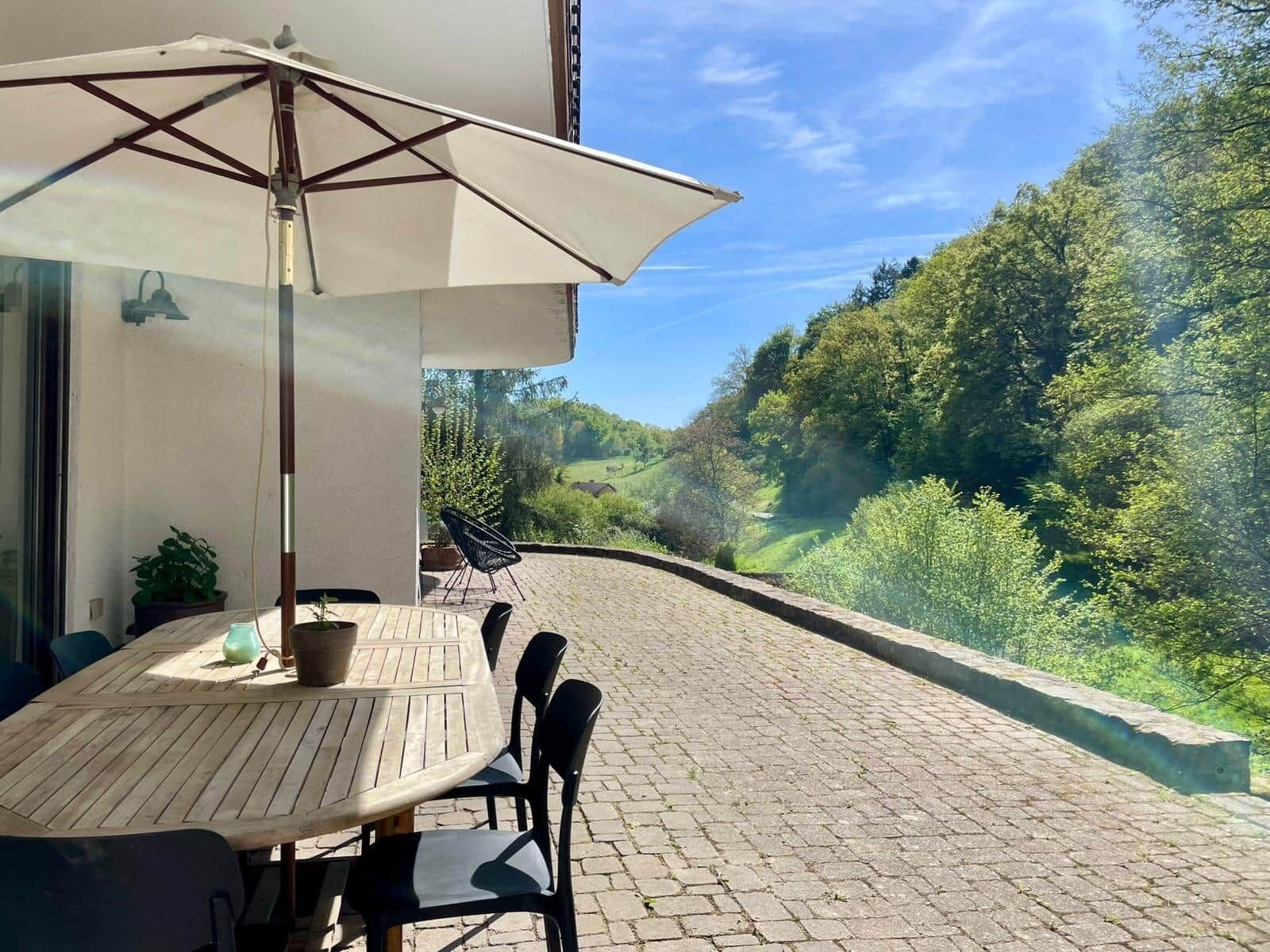 Eine Terrasse im Freien mit Tisch und Stühlen, einem großen Sonnenschirm und Blick auf einen grünen Hügel.