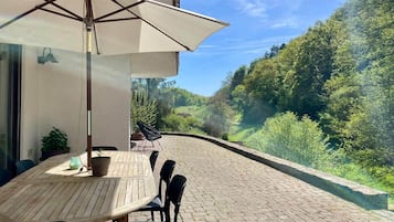 Eine Terrasse im Freien mit Tisch und Stühlen, einem großen Sonnenschirm und Blick auf einen grünen Hügel.