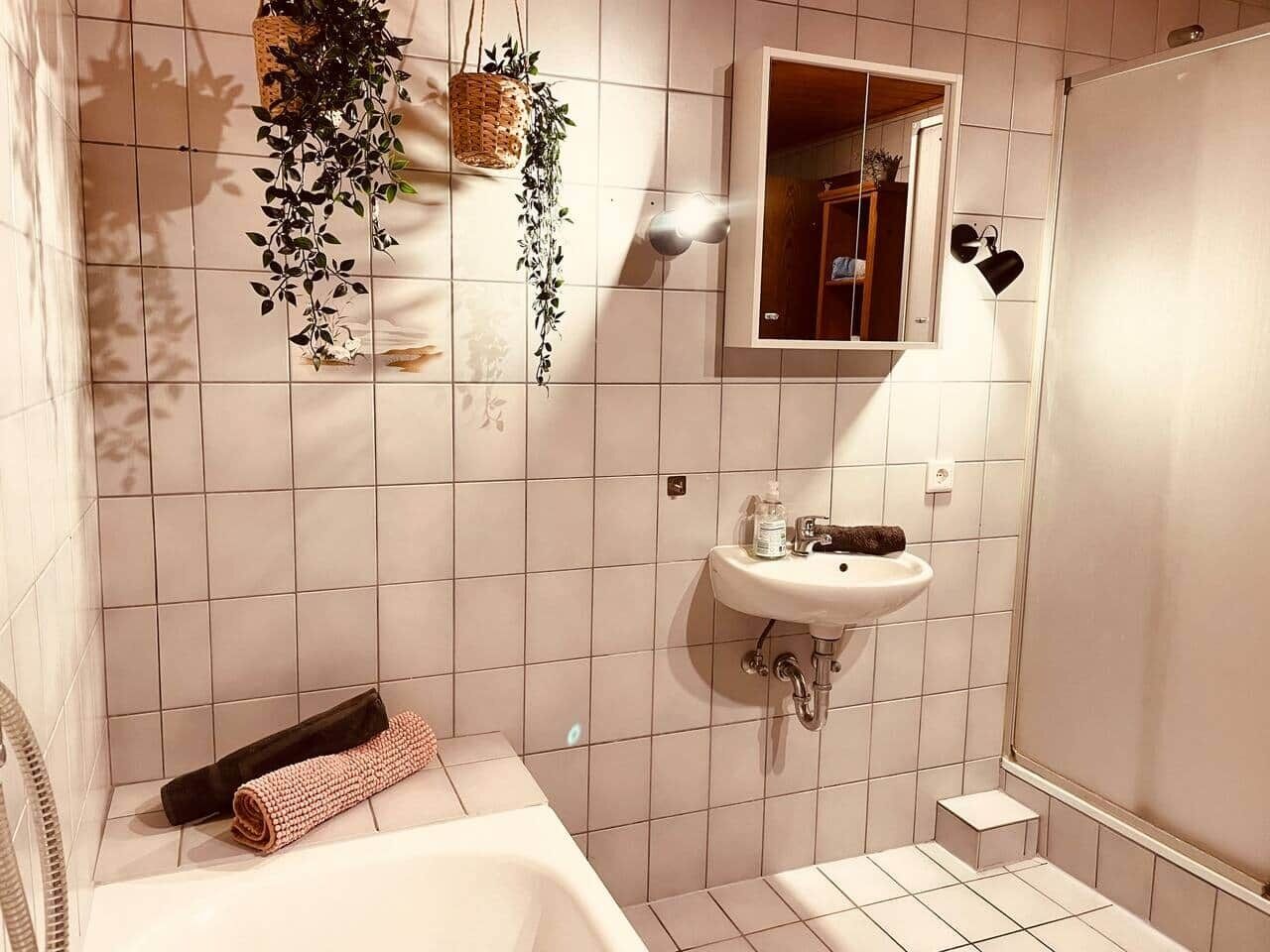 Un baño con una bañera blanca, un lavabo y un espejo. Hay plantas colgantes y una toalla en el suelo.