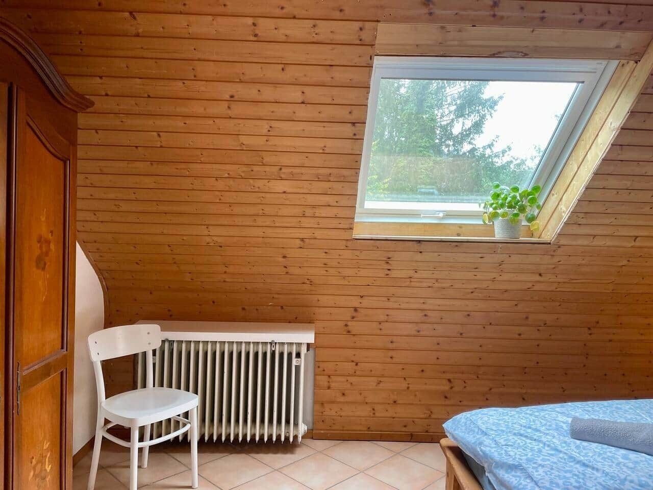 A small room with wooden walls, a radiator, a white chair, a bed with a blue cover, and a skylight with a plant.