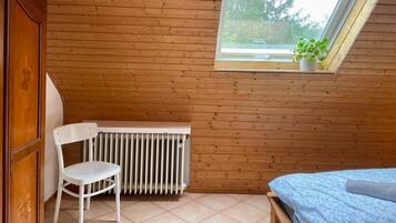A small room with wooden walls, a radiator, a white chair, a bed with a blue cover, and a skylight with a plant.