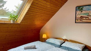 Een gezellige kamer met schuine wanden, houten lambrisering, een bed met beddengoed met een patroon, een bedlamp en een raam met een plant.
